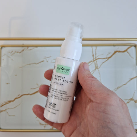 Hand holding a bottle of Mageau Magnesium Gentle Hand Lotion against a marble background